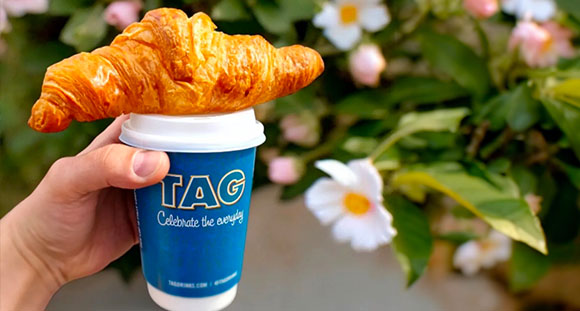 a person holding a coffee cup with a croissant balanced on top in front of a camellia bush